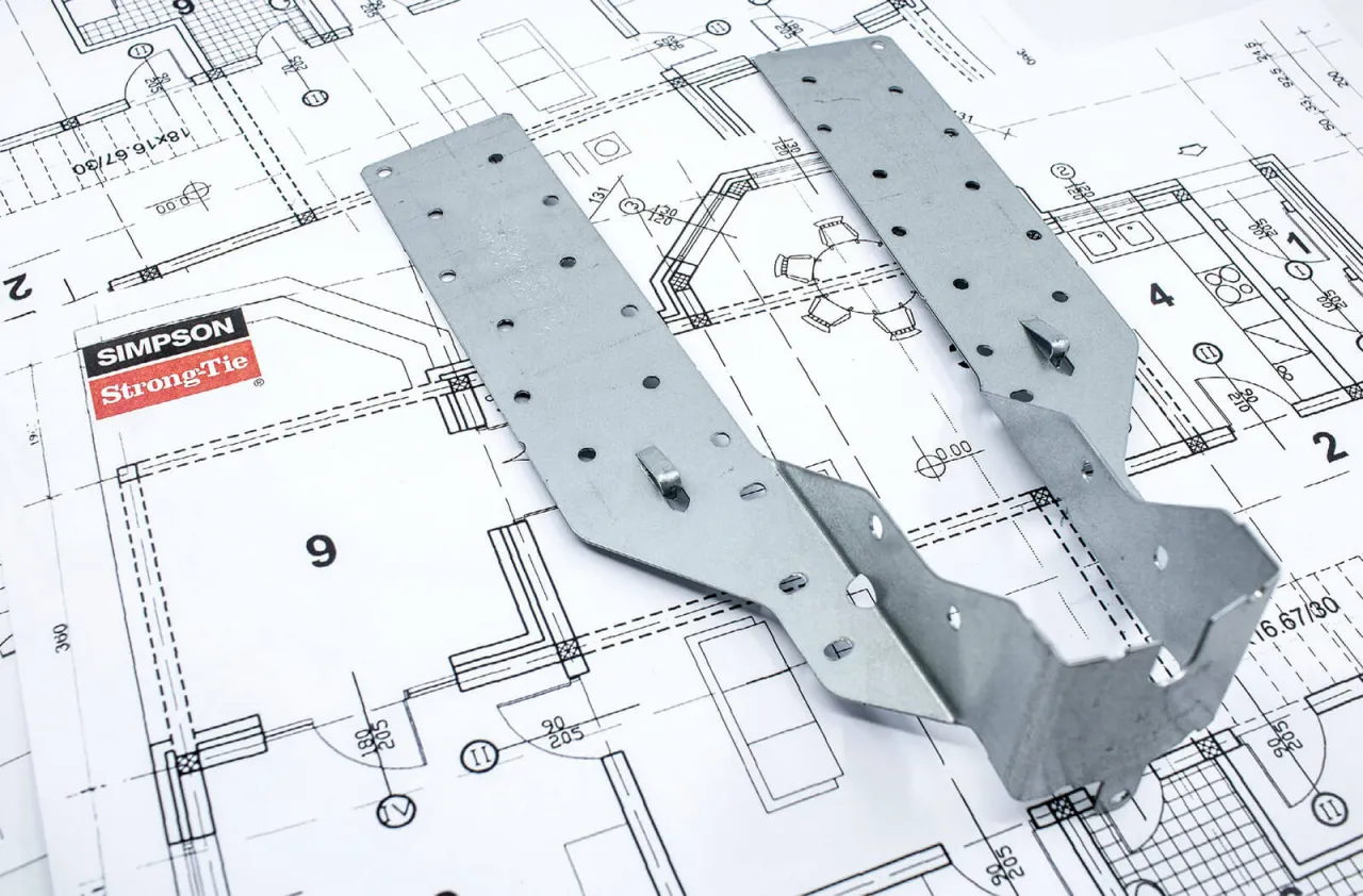 Metalowe łączniki Simpson Strong-Tie na tle plan&oacute;w architektonicznych. Pokazują, jak łączyć drewno konstrukcyjne, zapewniając stabilność i bezpieczeństwo budowy.