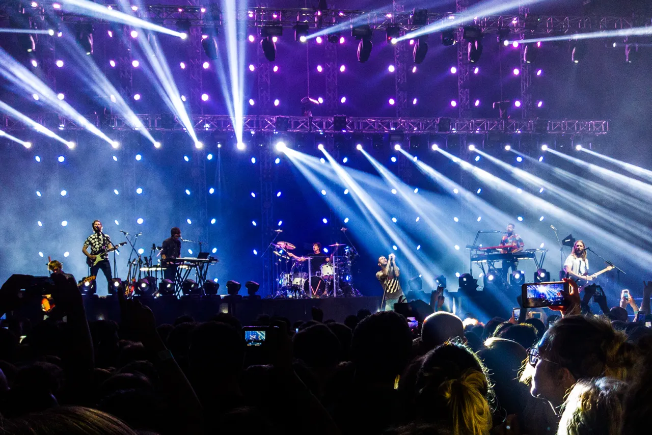 Koncert Maroon 5 w pełnym blasku świateł. Zesp&oacute;ł gra na scenie, a tłum bawi się w rytm muzyki.