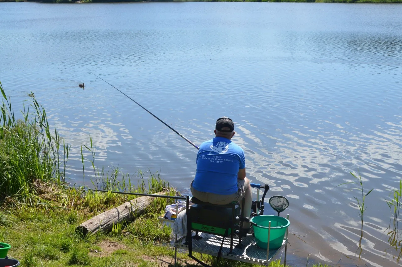 Wędkarz siedzi nad jeziorem, łowiąc ryby. Na brzegu widać zakaz używania zanęty.