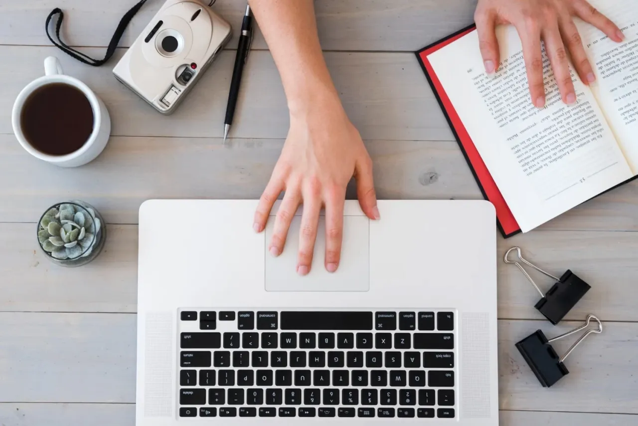 Ręce na laptopie, obok kawa, aparat, książka i roślina. Idealne miejsce, by dowiedzieć się, jak zacząć bloga.
