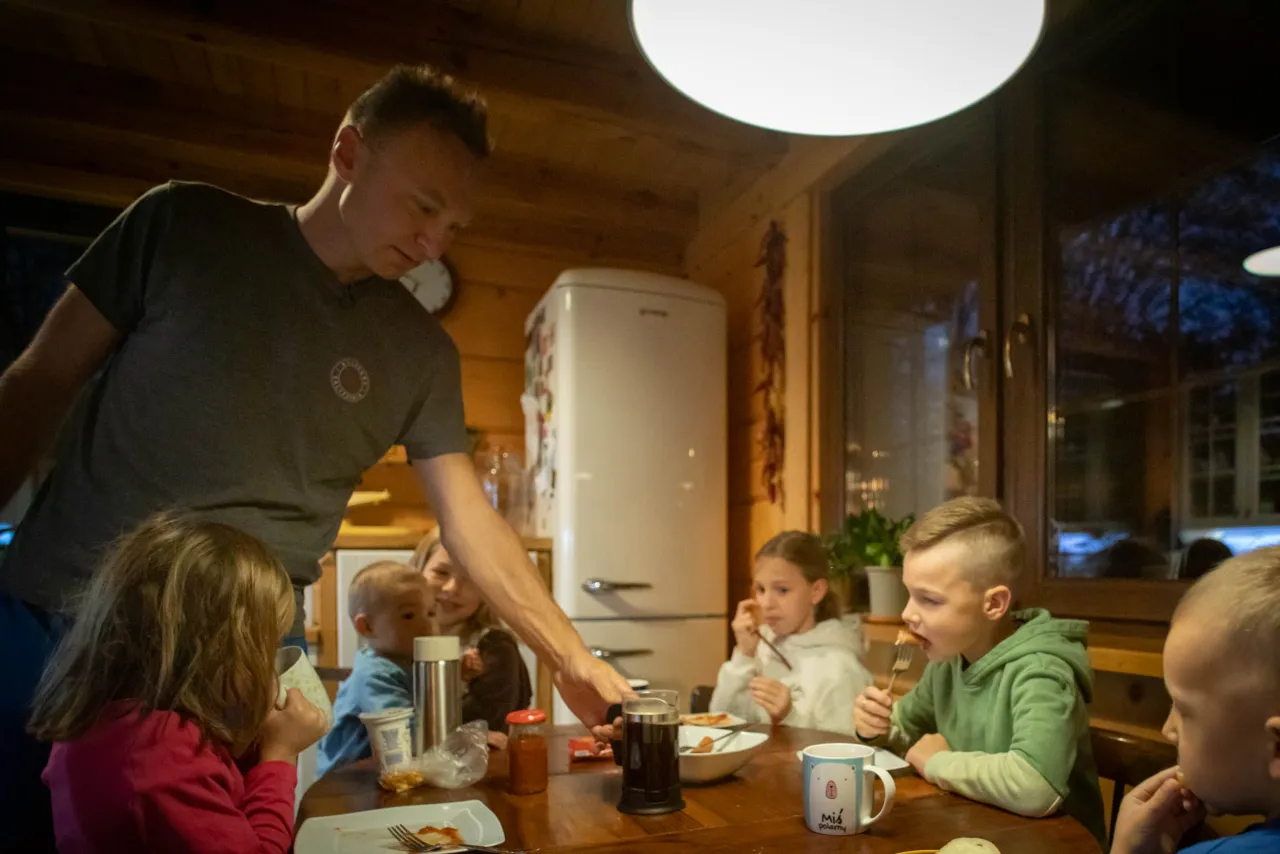 Ojciec z dziećmi przy stole, gdy matka zostawia dziecko ojcu. Rodzina je kolację w przytulnym, drewnianym wnętrzu.