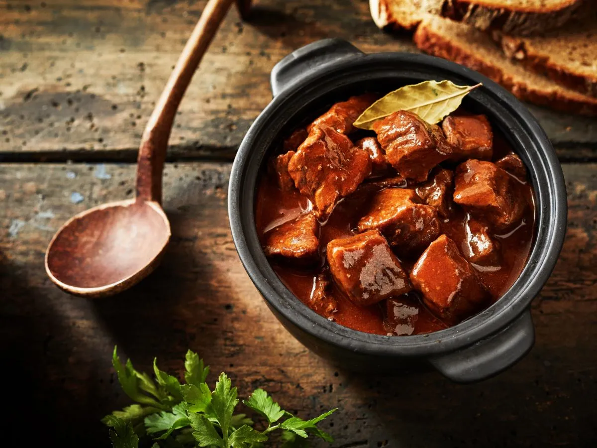 Gulasz w garnku, obok drewniana łyżka i kromki chleba. Idealna porcja mięsa na osobę na obiad.