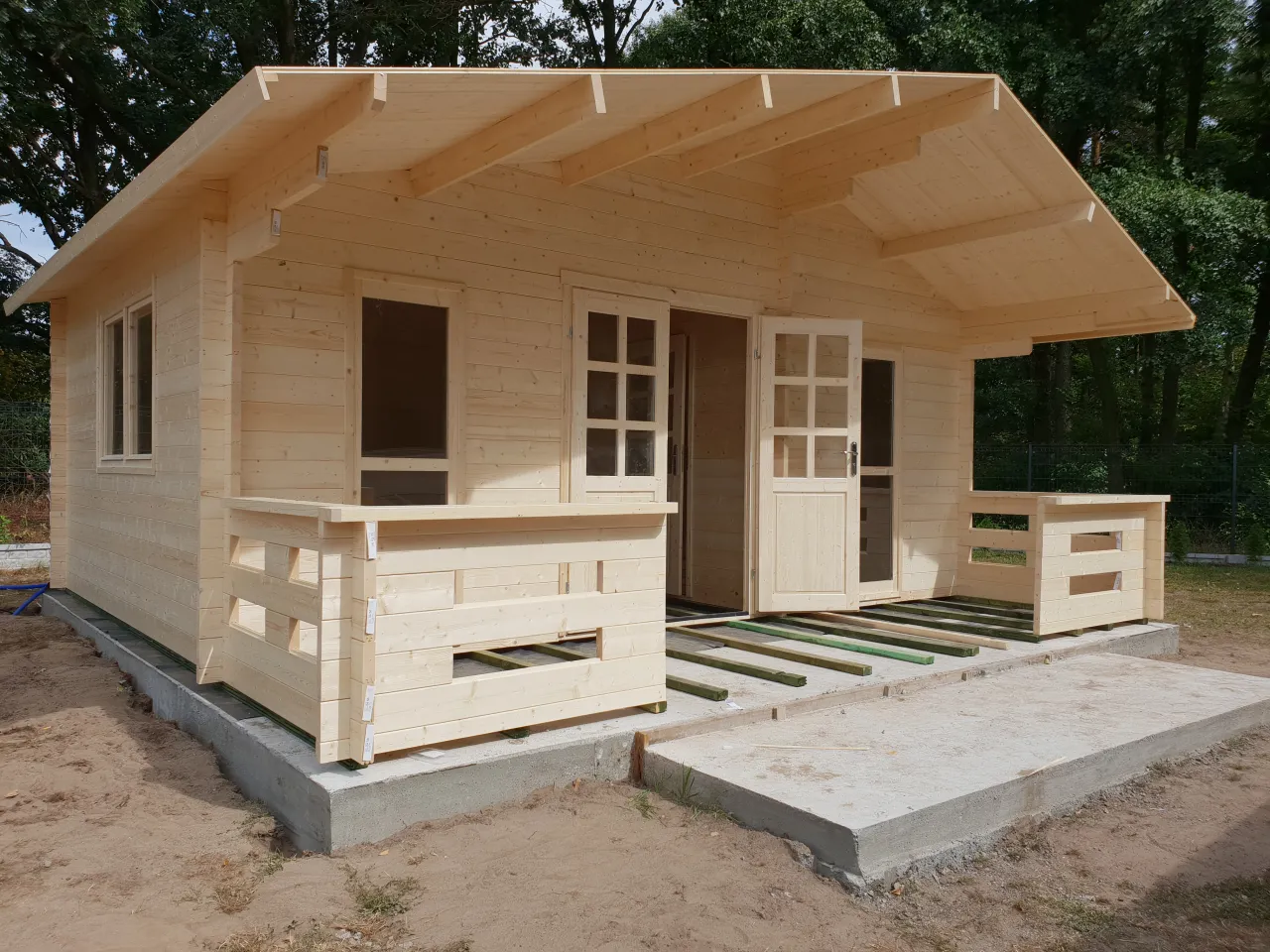 Nowy, drewniany domek ogrodowy z werandą i otwartymi drzwiami, gotowy na przyjęcie pierwszych gości.