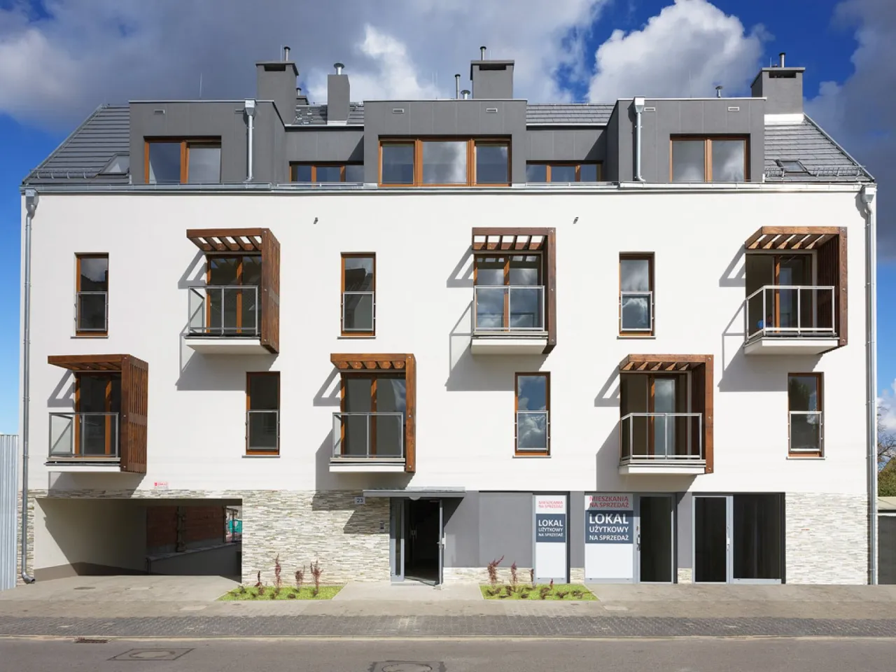 Nowoczesne osiedle z białymi balkonami i drewnianymi elementami. Widać wejście do budynku i szyldy 