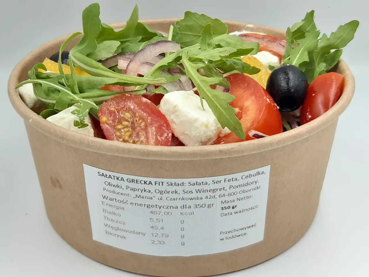 Sałatka grecka w pudełku z etykietą informującą, ile kalorii ma sałatka grecka: 467 kcal na 350g. Widoczne pomidory, ser feta, oliwki, rukola.