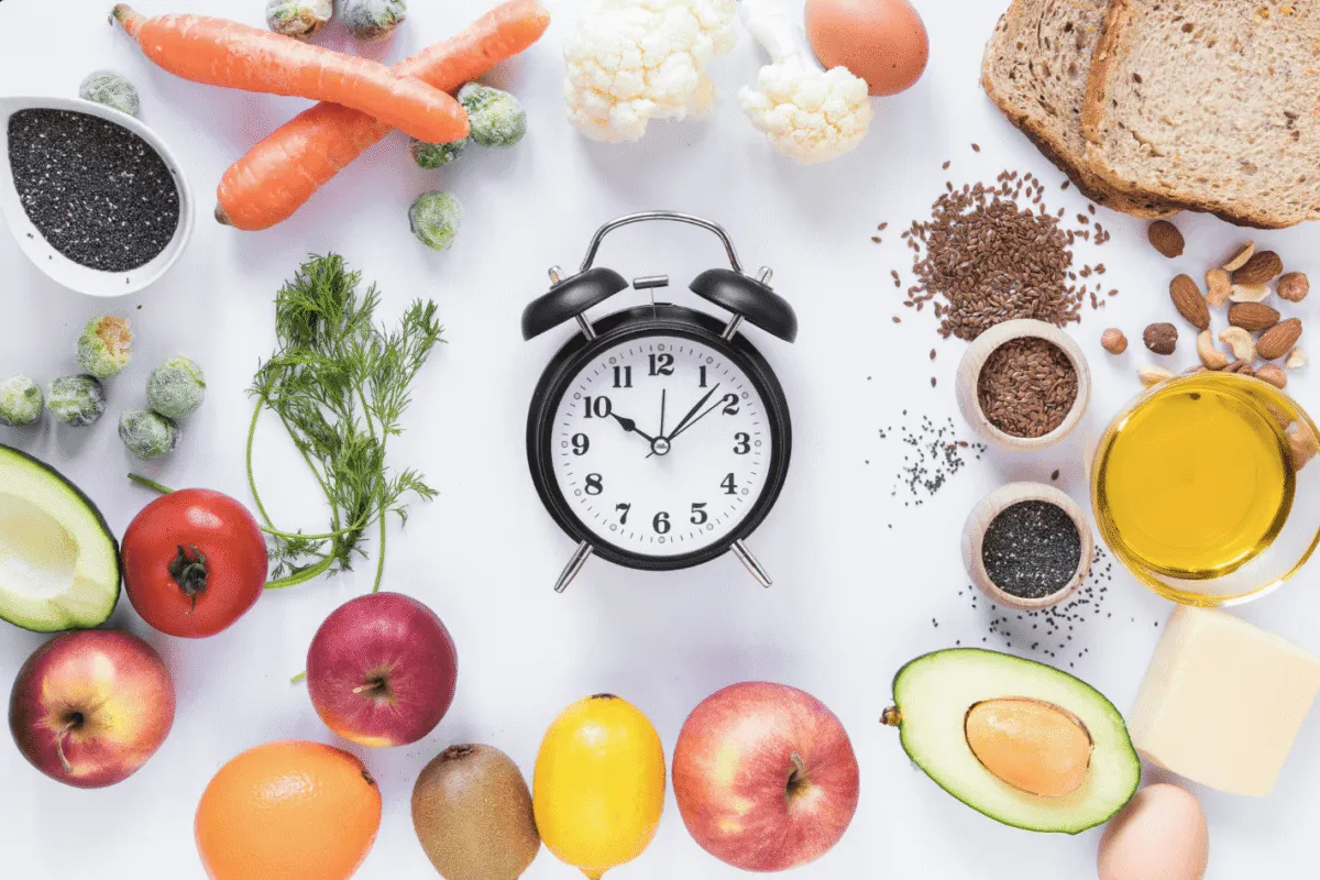 Zegar i zdrowe produkty: marchew, jabłka, awokado, pomidor, cytryna, kiwi, jajko, oliwa, orzechy, nasiona, chleb. Klucz do tego, co ile jeść, żeby schudnąć.