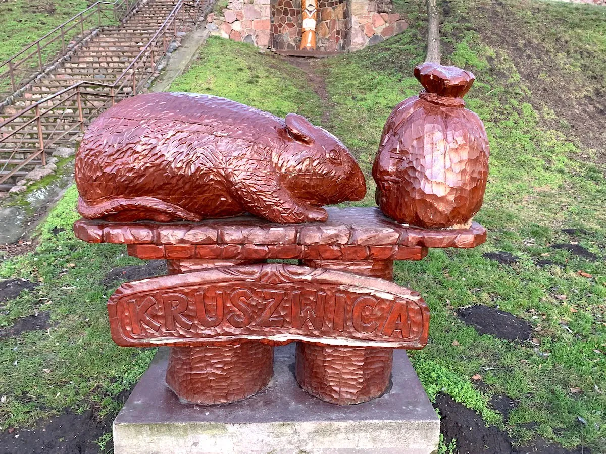 Drewniana rzeźba szczura i worka, nawiązująca do baśni o Popielu. Podpis