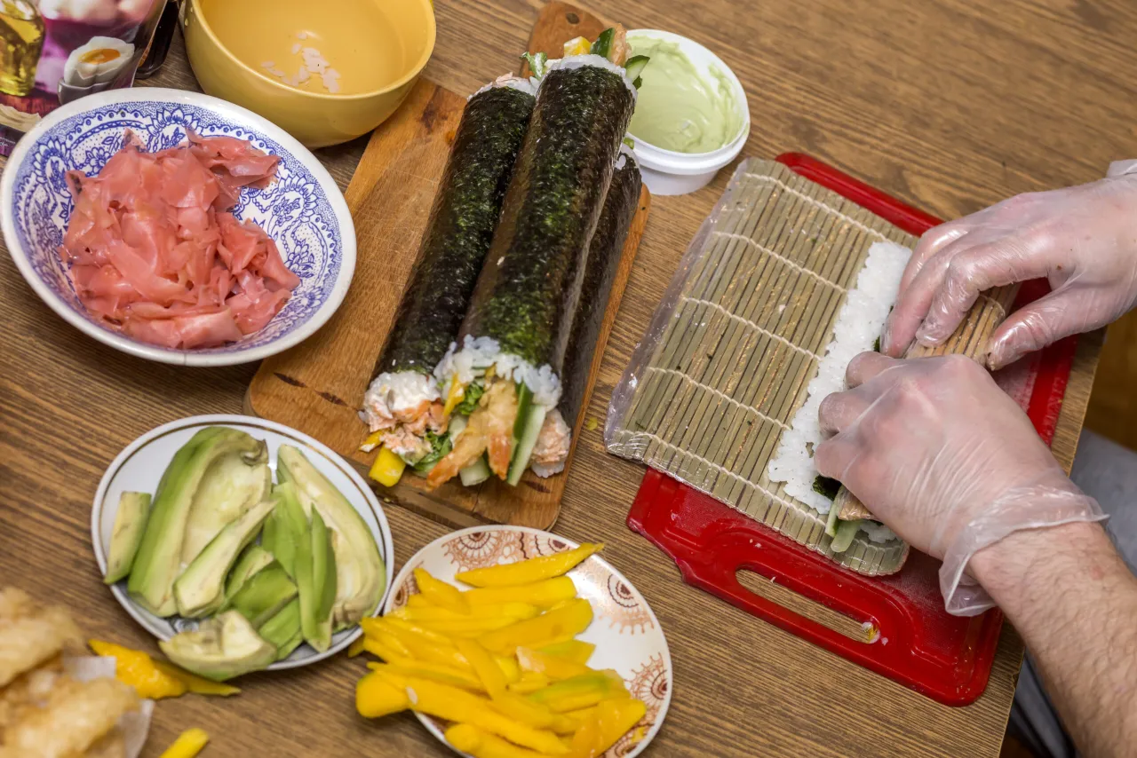 Ręce w rękawiczkach zwijają sushi na macie. Na stole składniki: ryż, nori, awokado, mango, imbir i sos.