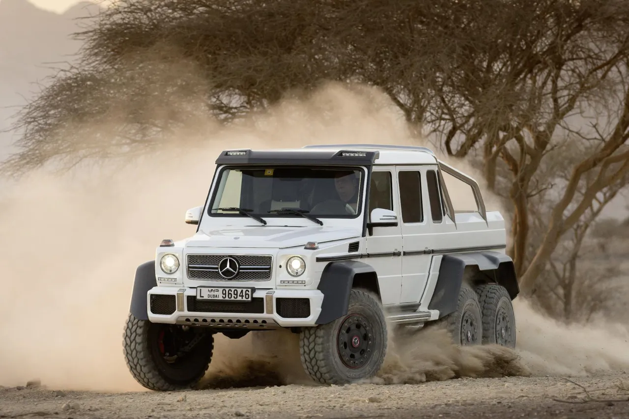 Biały Mercedes-Benz G63 AMG 6x6 szaleje po pustyni, wzbijając tumany kurzu.