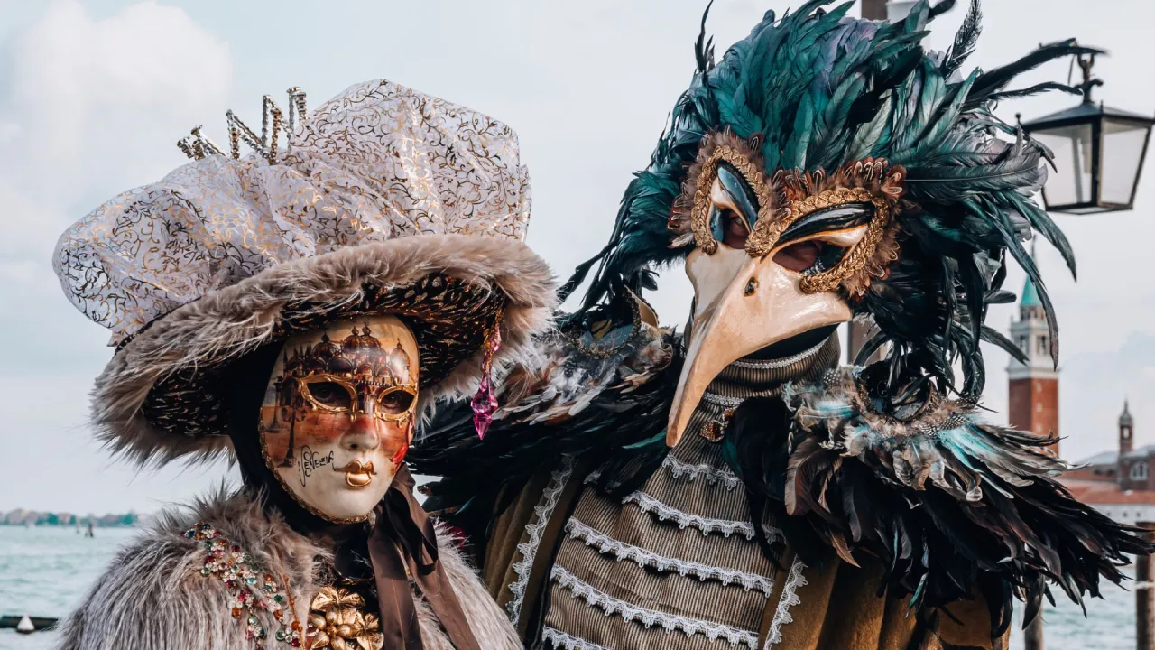Dwie postacie w maskach weneckich, jedna z ptasim dziobem, druga ozdobiona złotymi wzorami. Maski weneckie co oznaczają? Tajemnicę i zabawę.