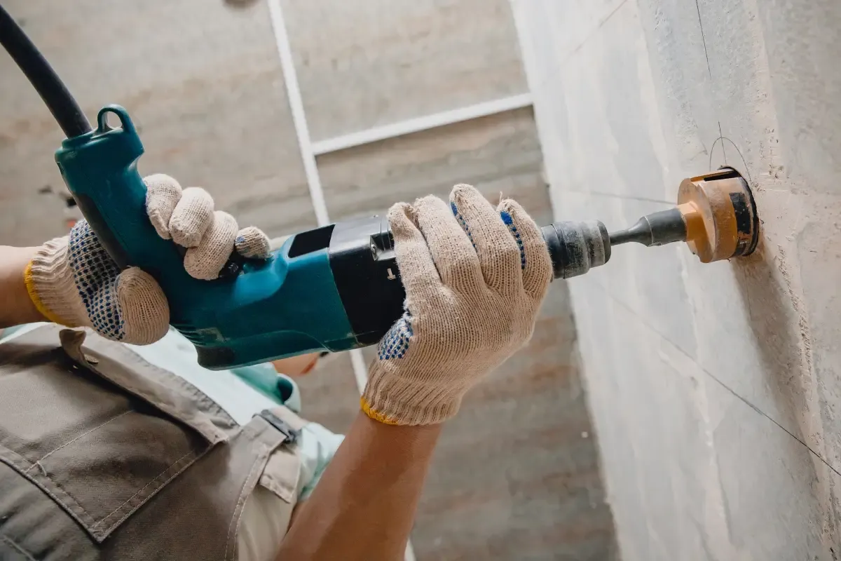 Ręce w rękawicach trzymają wiertarkę z otwornicą, wiercąc otw&oacute;r w ścianie. To pierwszy krok, jak zrobić bruzdy pod kable.