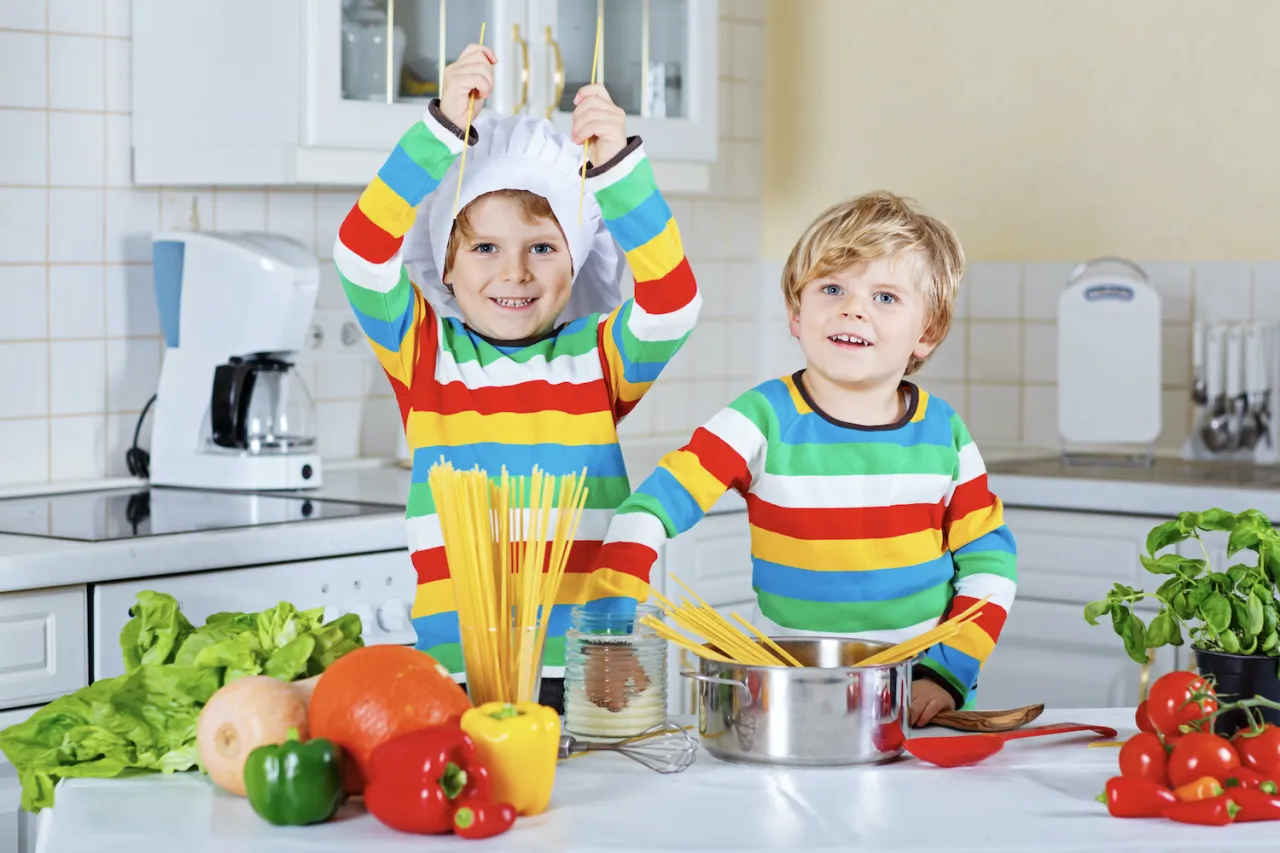 Dw&oacute;ch chłopc&oacute;w w kolorowych bluzach i czepkach kucharskich przygotowuje spaghetti. Widać warzywa i makaron. Idealne danie, co może ugotować 10 latek.