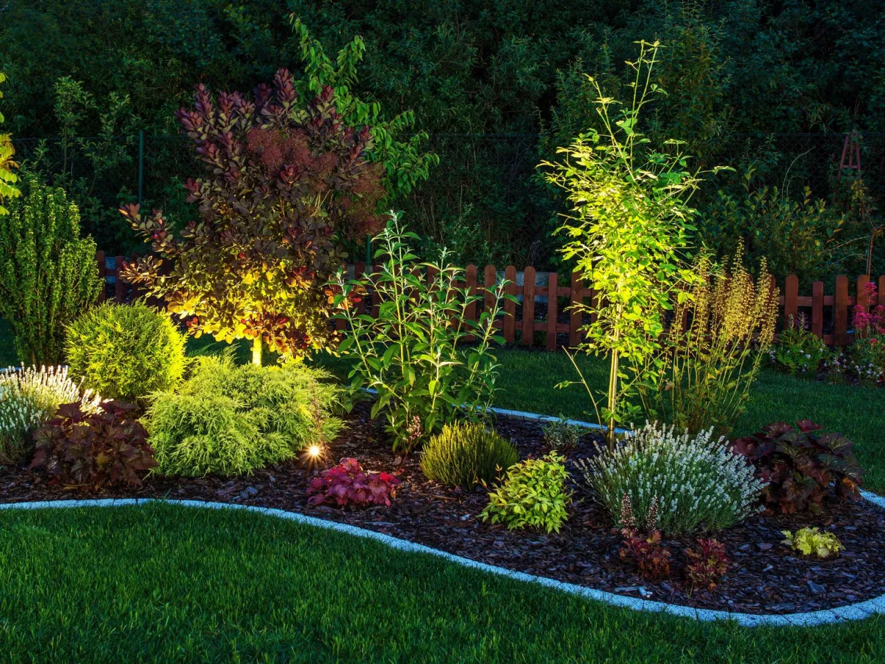 Oświetlony ogr&oacute;d przed domem, pełen zieleni i kolorowych roślin. Inspiracja, jak zaprojektować ogr&oacute;d przed domem, by zachwycał nocą.