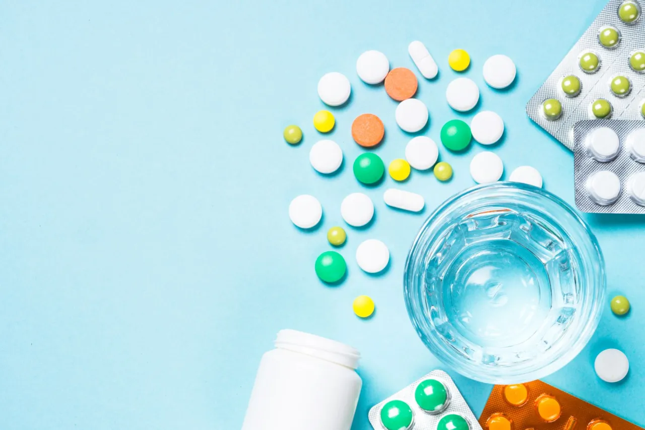 Leki dla senior&oacute;w 65 plus: białe, zielone i pomarańczowe tabletki, blistry, szklanka wody i biały pojemnik na leki.