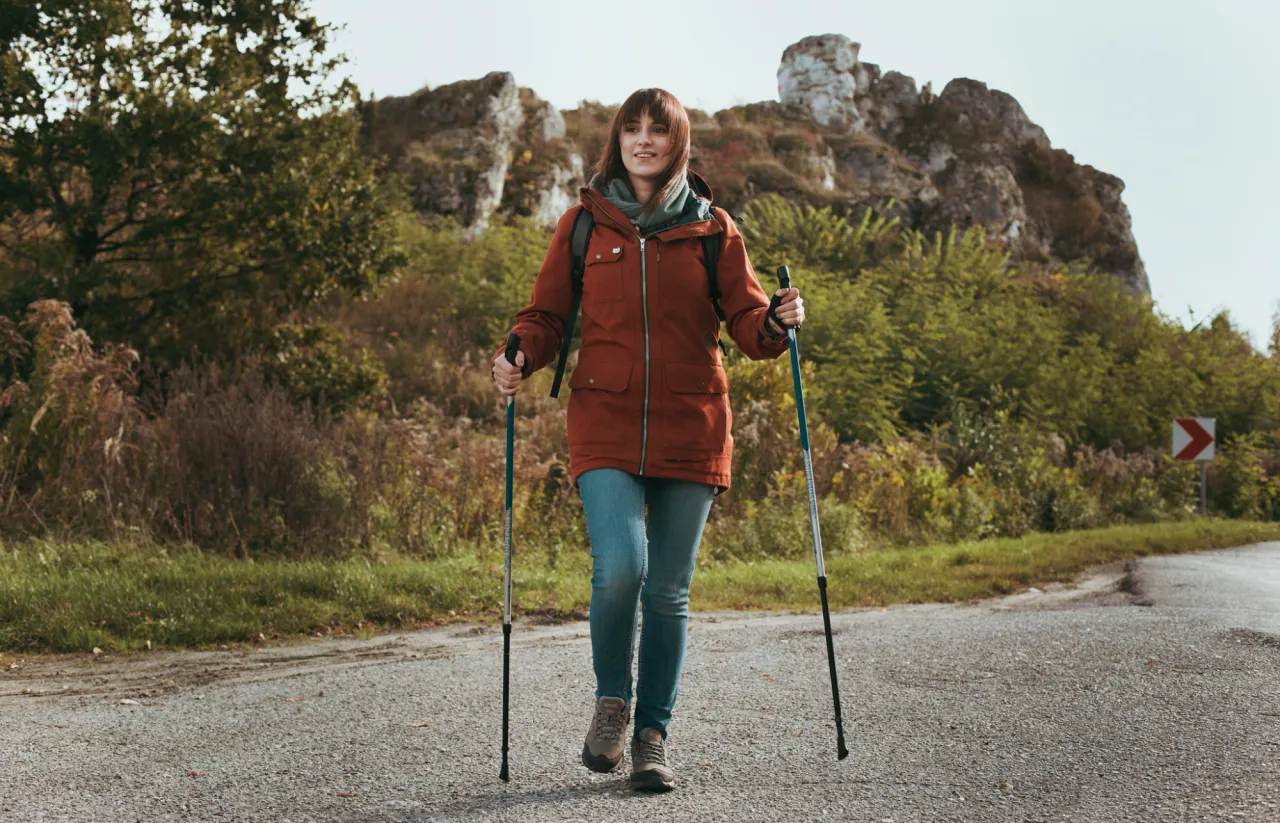 Kobieta z kijkami w g&oacute;ry idzie po drodze. W tle skaliste wzg&oacute;rza i jesienna roślinność.