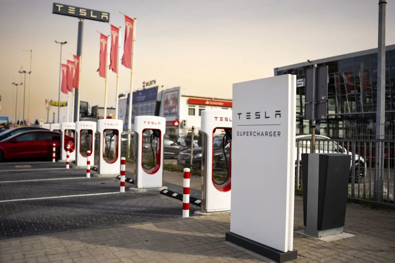 Ładowanie Tesli w domu może być tańsze niż na stacji. Widok na stację Supercharger Tesli z czerwonym samochodem w tle.