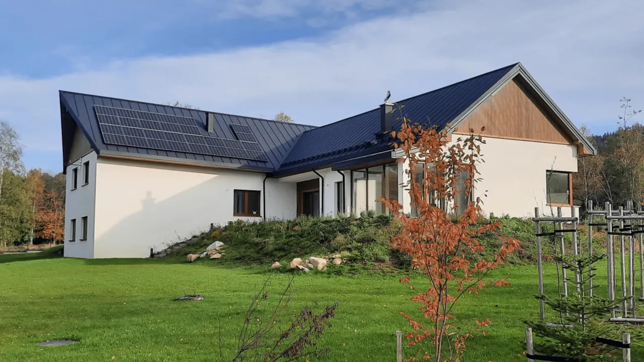 Nowoczesny ekologiczny dom z panelami słonecznymi na dachu, otoczony zielonym ogrodem i jesiennymi drzewami.