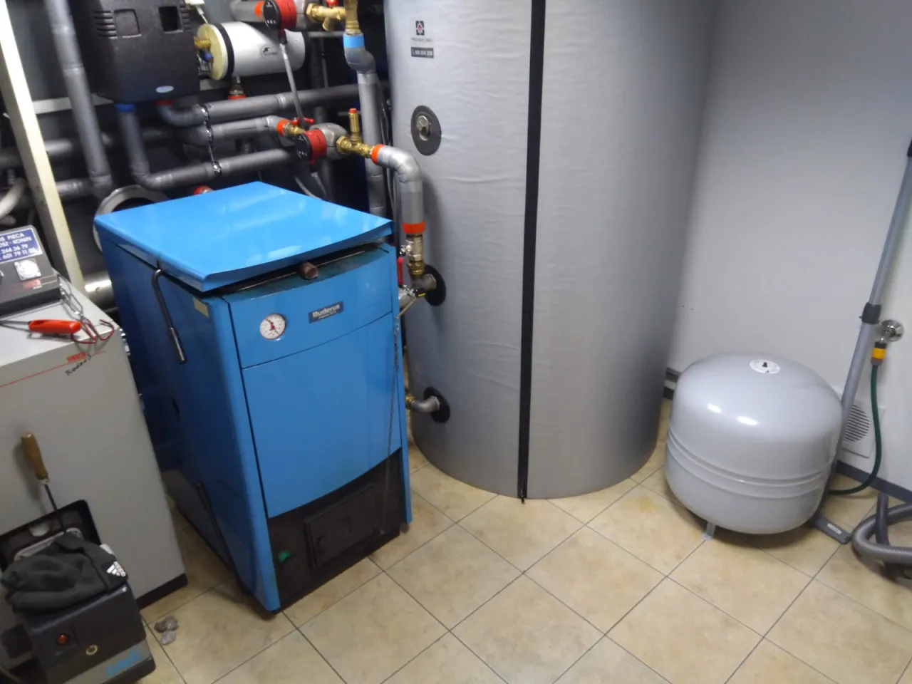 Niebieski kocioł co na drewno Buderus obok dużego zasobnika wody i zbiornika przeponowego.