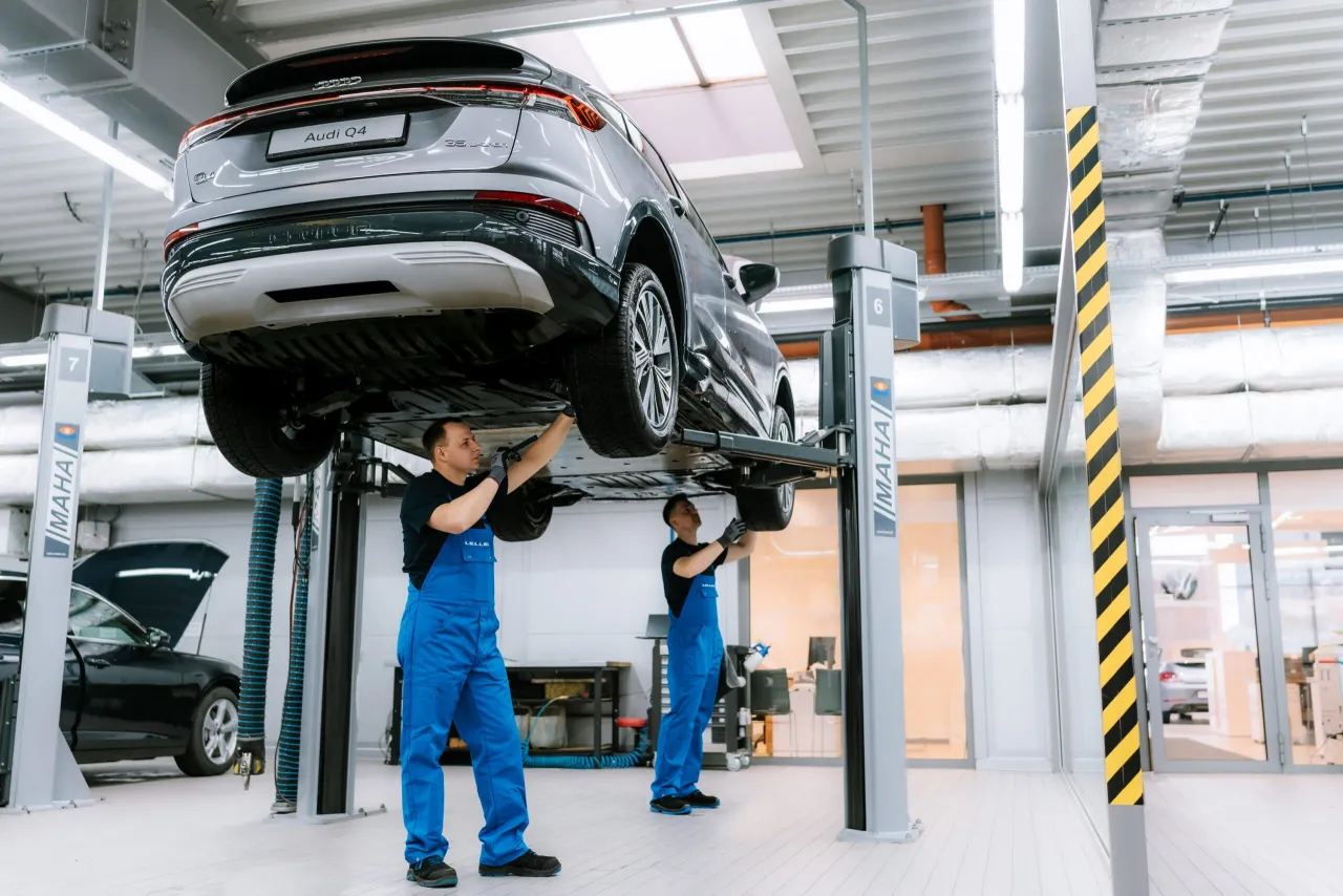 Mechanicy w warsztacie podnoszą Audi Q4 e-tron na podnośniku. Jeden z nich sprawdza podwozie, drugi pracuje przy kole.
