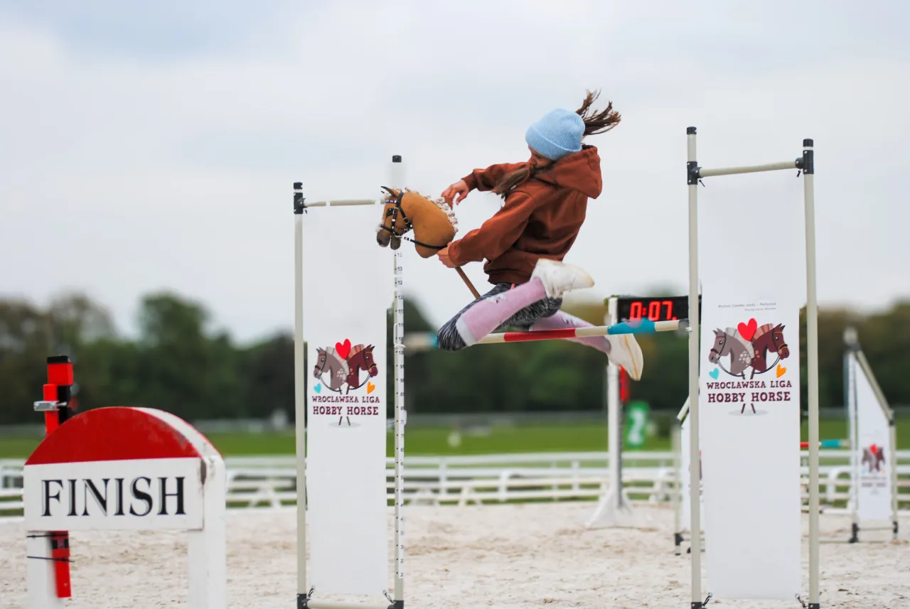 Dziewczynka skacze przez przeszkodę na kucyku. To zawody w stylu hobby horse, gdzie liczy się zwinność i precyzja.