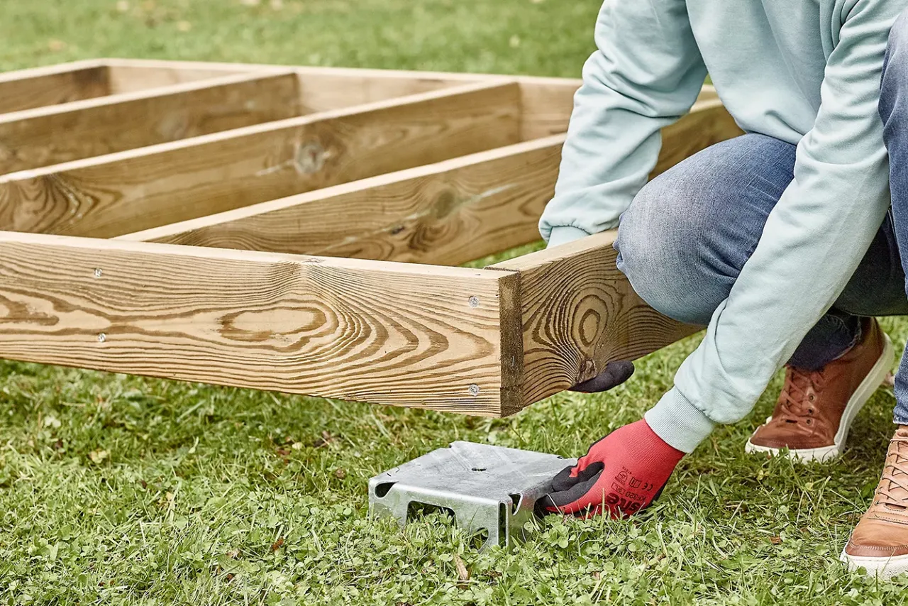 Osoba w rękawiczkach montuje drewnianą konstrukcję, która będzie podstawą dla domku narzędziowego. Podpora z bloczków betonowych czeka na swoje miejsce.