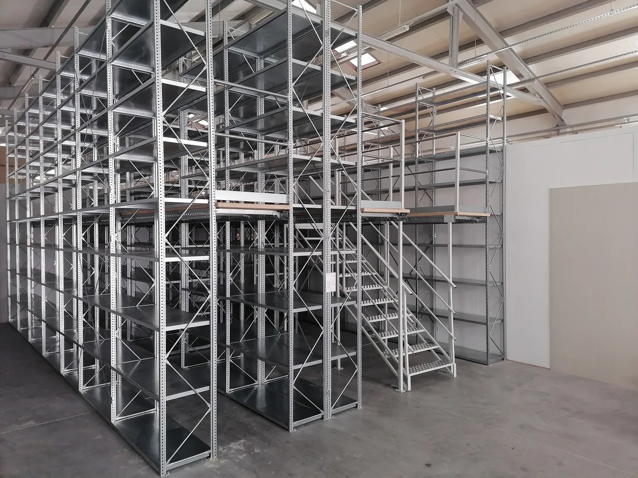 W magazynie widać wielopoziomowe regały z metalowymi belkami, tworzące antresolę. Schody prowadzą na wyższe piętro.