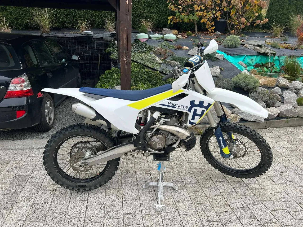 Biały motocykl cross Husqvarna 250 na stojaku, gotowy do jazdy pośród jesiennego ogrodu z oczkiem wodnym.