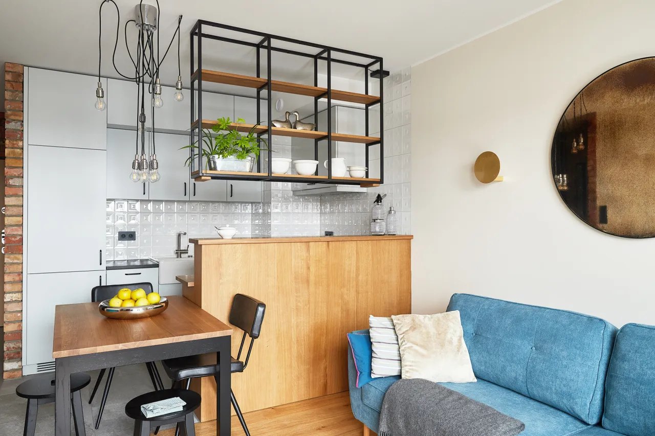 Stylowa aranżacja małego mieszkania: kuchnia z drewnianym blatem i niebieska kanapa. Industrialne półki i designerska lampa dodają charakteru.