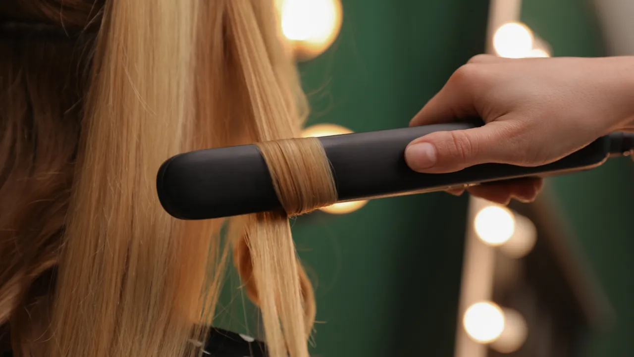 Dłoń modeluje blond włosy prostownicą, tworząc loki. Poznaj triki, jak utrwalić loki z prostownicy, by fryzura trzymała się cały dzień.