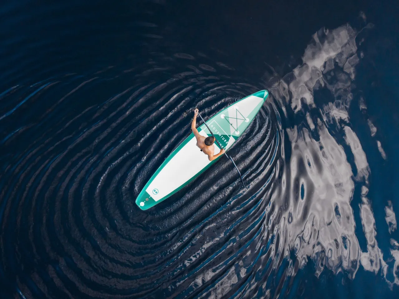 Mężczyzna wiosłuje na dmuchanej desce do pływania po spokojnej wodzie, tworząc kręgi na powierzchni.