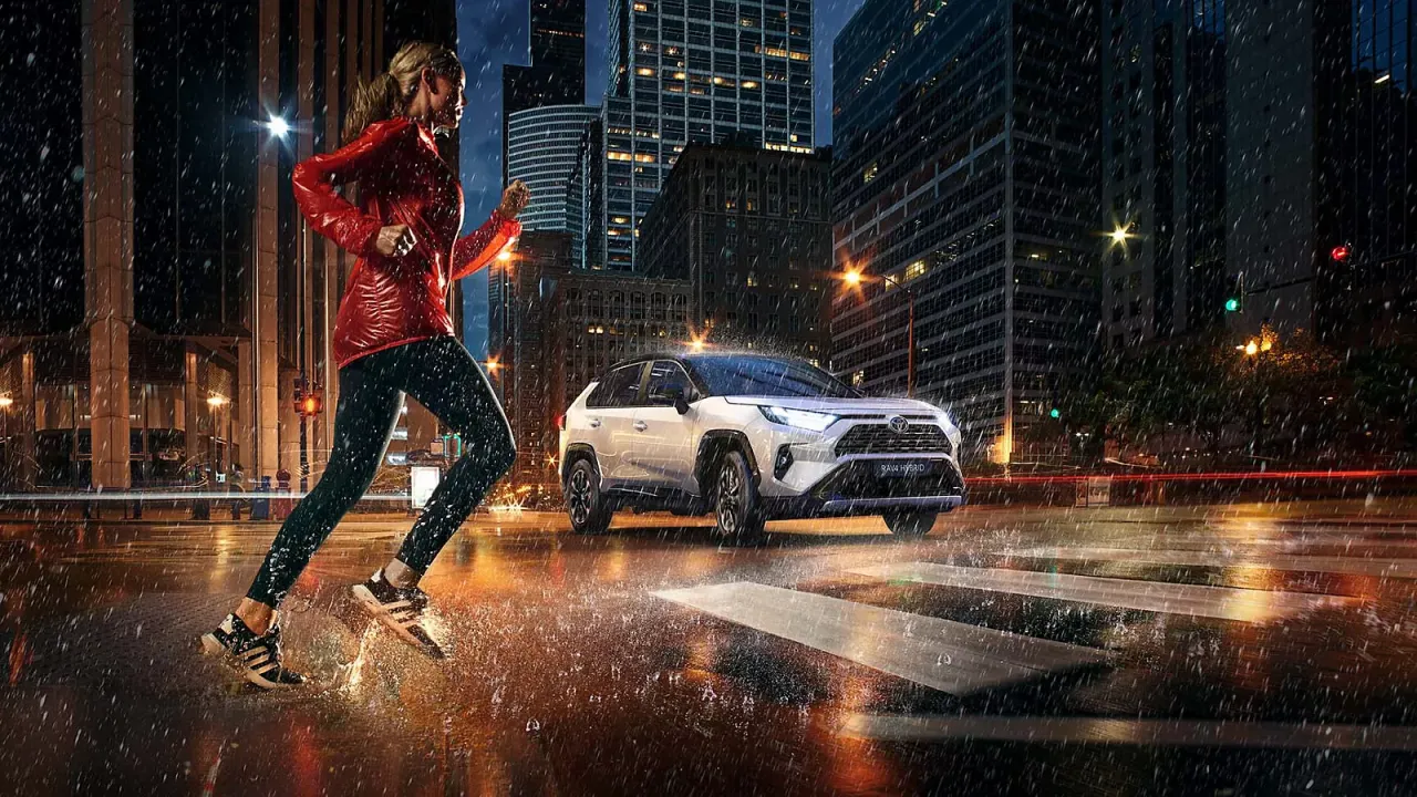 Kobieta biegnie w deszczu po mieście, obok niej jedzie biała Toyota RAV4 Hybrid. Zastanawiasz się, ile pali Toyota RAV4 Hybrid?