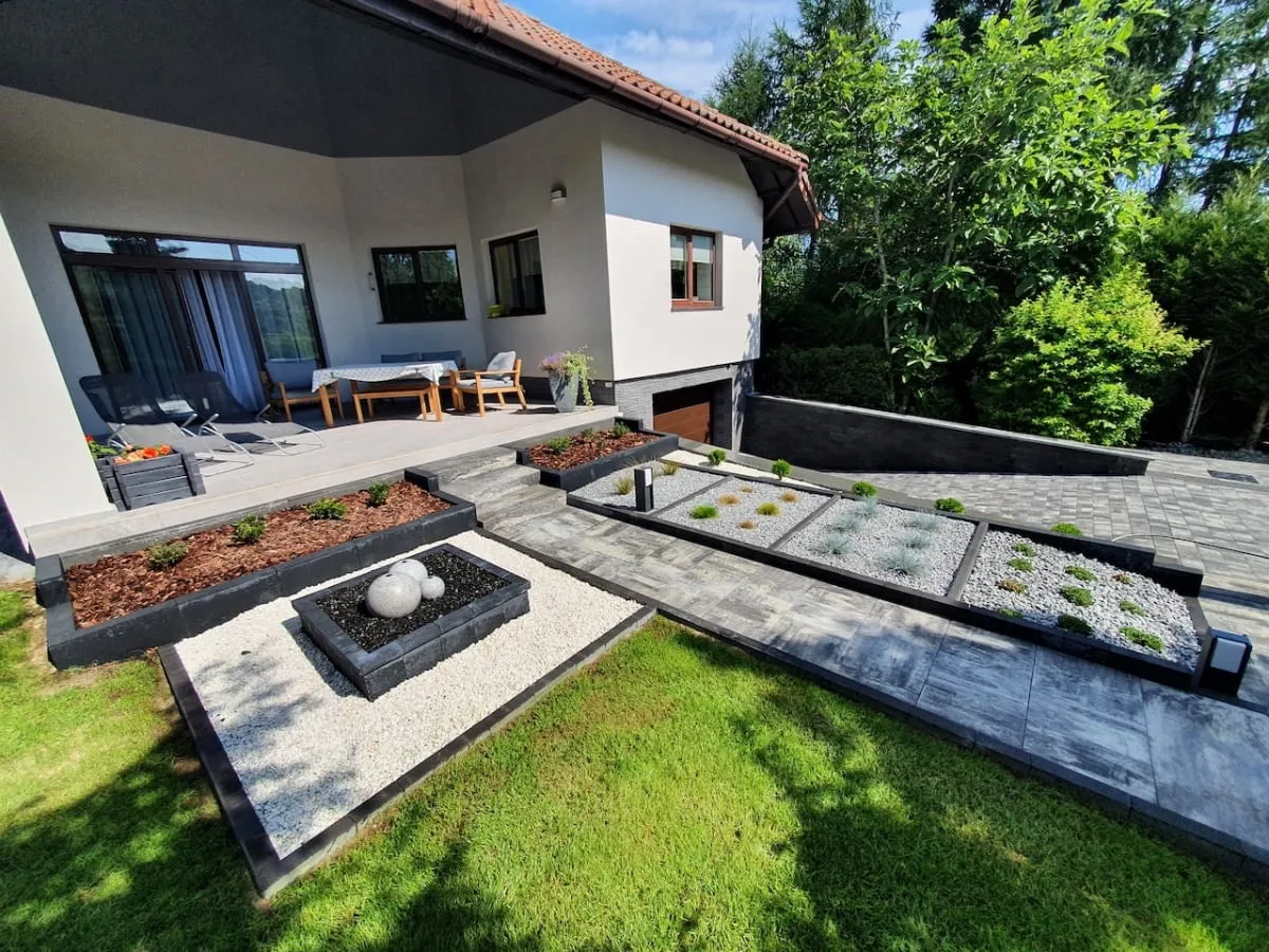 Nowoczesny podjazd z kostki brukowej, rabaty z kamieniami i roślinami, idealne rozwiązanie co na utwardzenie podjazdu.