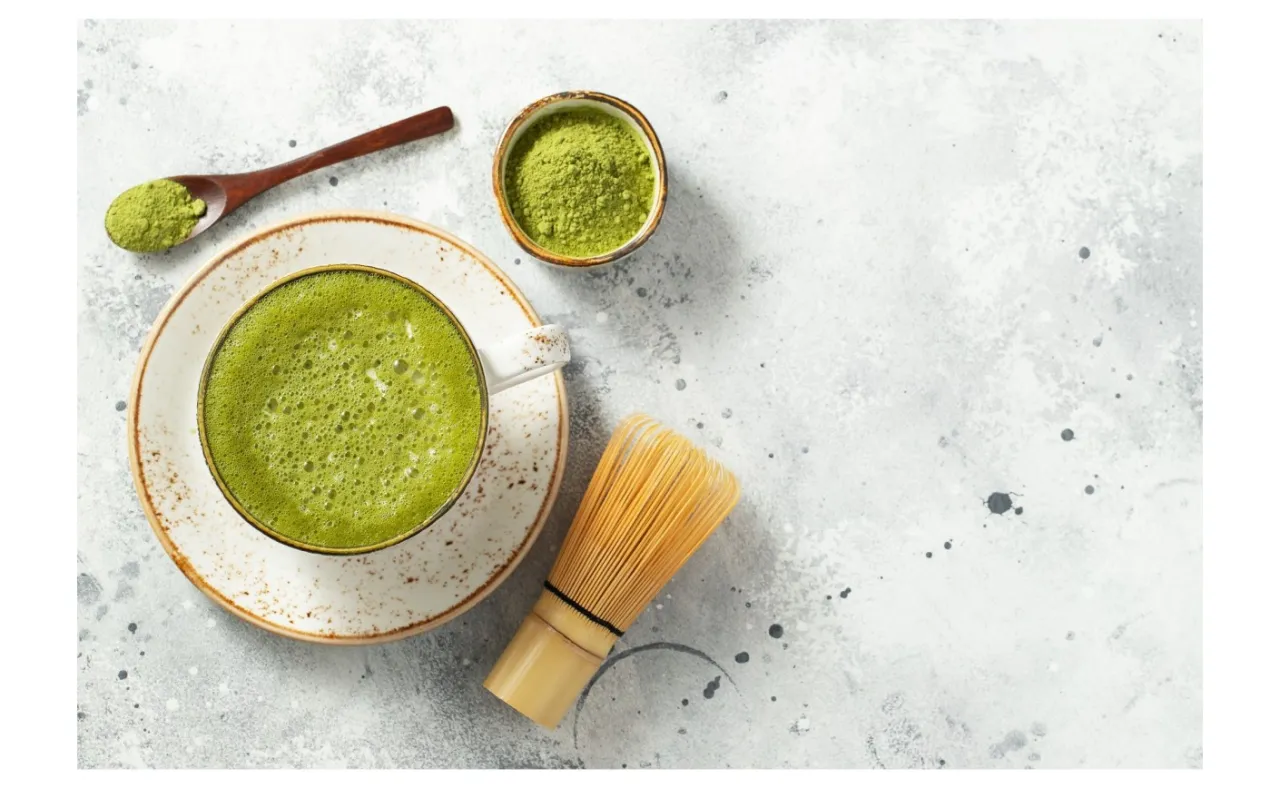 Zielona, spieniona matcha w filiżance, obok bambusowa miotełka i łyżeczka z proszkiem. Idealne akcesoria, by dowiedzieć się, jak pić matcha.
