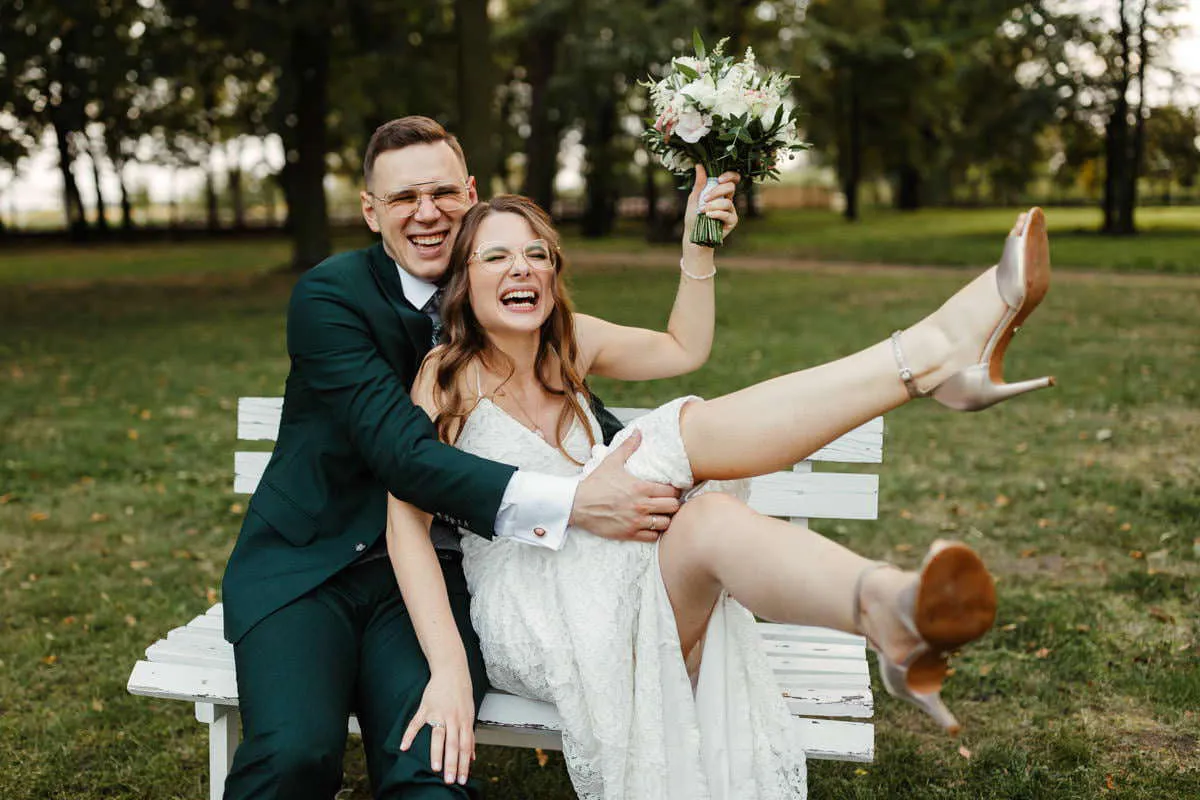 Szczęśliwa para młoda na ławce, panna młoda z bukietem śmieje się radośnie. Idealny przykład, co napisać w mailu do fotografa ślubnego, by uchwycił takie emocje.