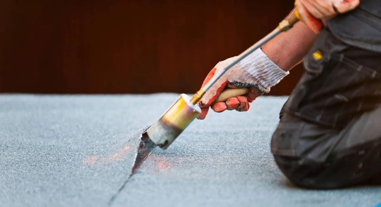 Pracownik w rękawicach z palnikiem zgrzewa papę na dachu, pokazując, jak naprawić dach pokryty papą.