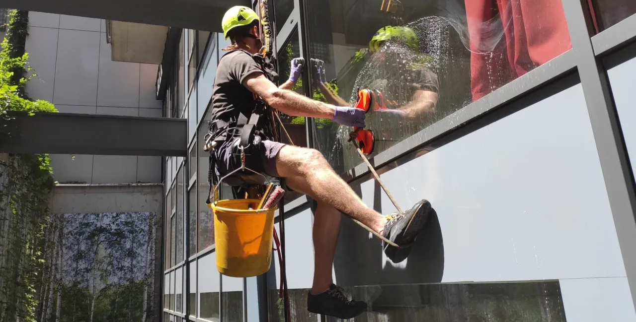 Specjalista wspinaczkowy myje okna na wysokości, używając sprzętu alpinistycznego. Pokazuje, jak myć okna na wysokości bezpiecznie i efektywnie.