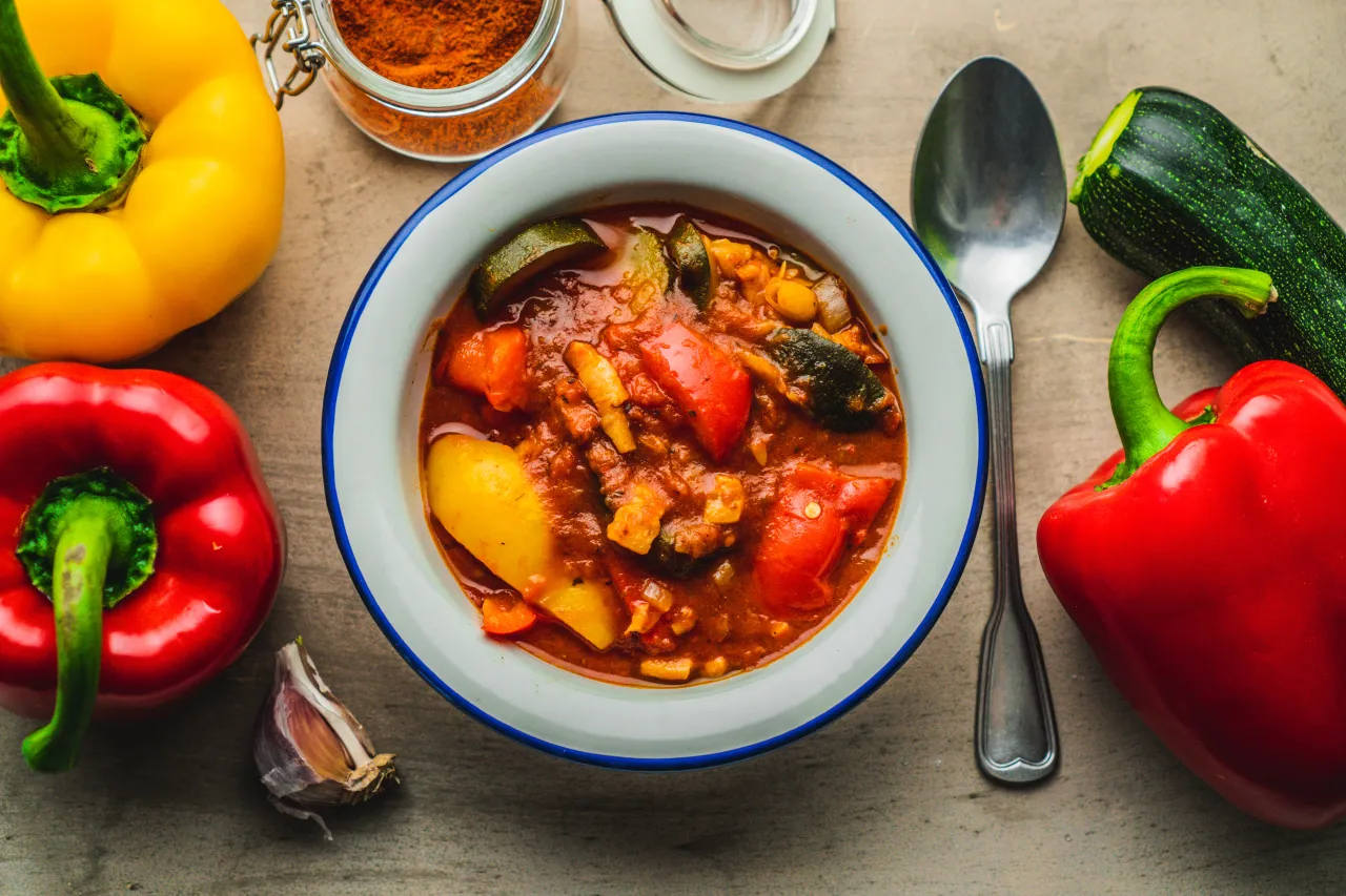 Gotowe leczo w misce, obok papryki, cukinii, czosnku i słoiczek z papryką. Idealne przyprawy do leczo to podstawa!