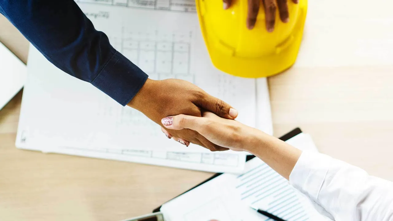 Uścisk dłoni na tle planów budowlanych i żółtego kasku. To symboliczne przedstawienie tego, co potrzebne do pozwolenia na budowę: współpraca i dokumentacja.