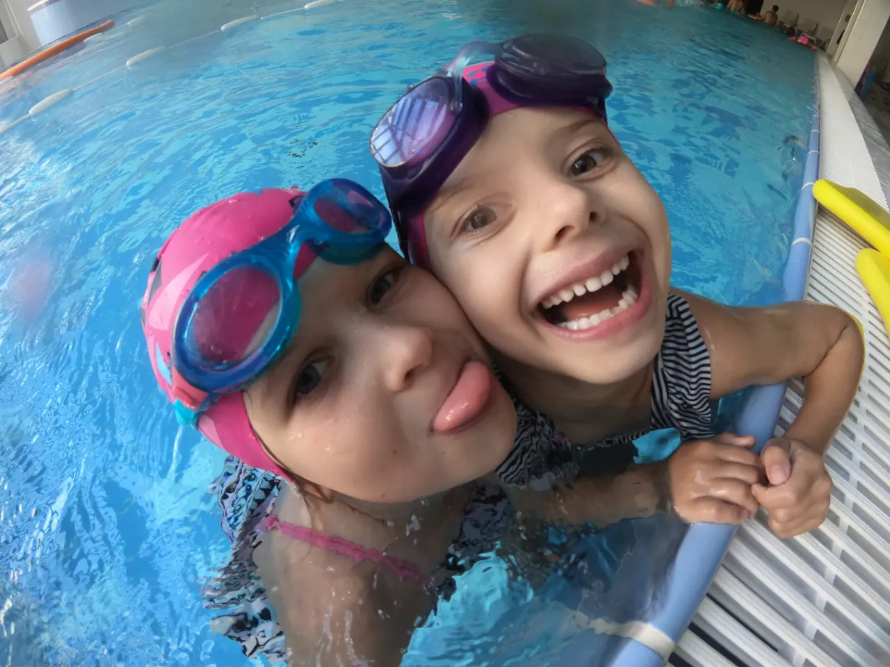 Dwie dziewczynki w czepkach i okularach pływackich w basenie. Jedna śmieje się, druga wystawia język. Nauka pływania to świetna zabawa!