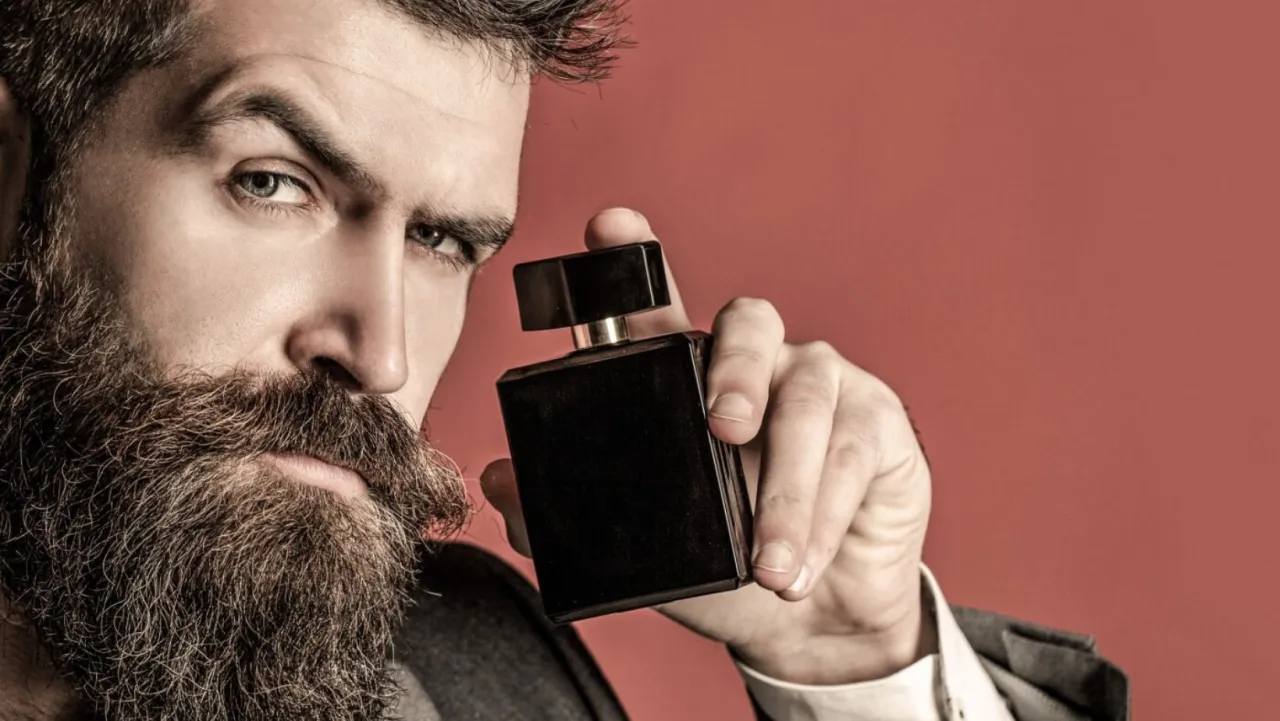 Mężczyzna z brodą trzyma czarną butelkę wody toaletowej. Dowiedz się, jak używać wody toaletowej, by podkreślić swój styl.