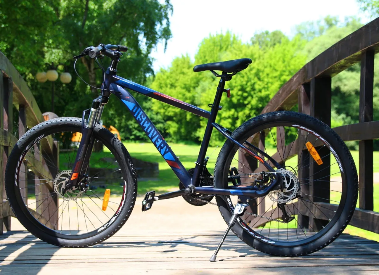 Ciemnoniebieski rower g&oacute;rski Discovery z pomarańczowymi i czerwonymi akcentami, idealny jako rower dla 13 latka, stoi na drewnianym moście.