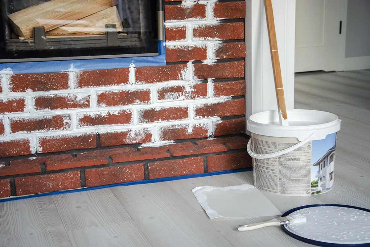 Malowanie obudowy kominka na biało. Wiadro z farbą i pędzel czekają, by nadać cegłom nowy wygląd.