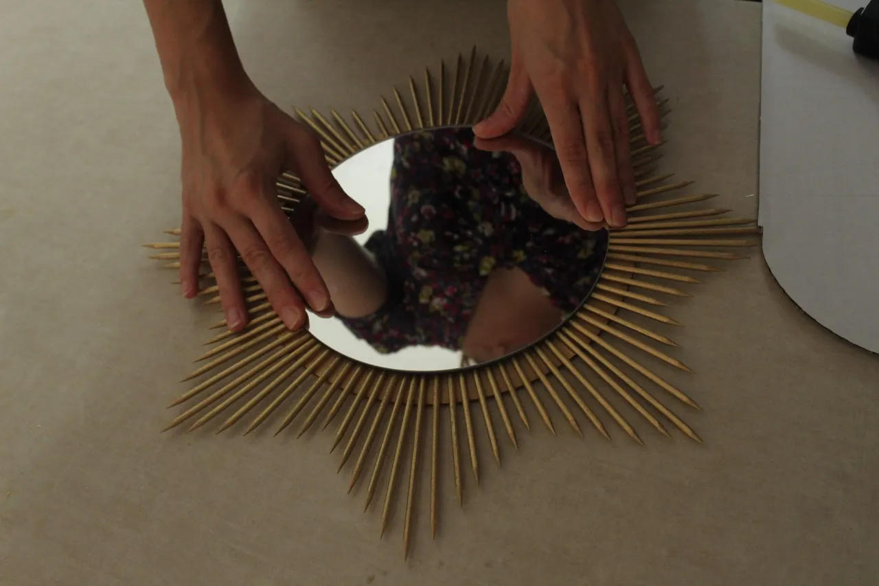 Okrągłe lustro w złotej ramie z patyczków, jak zrobić samemu. Dwie dłonie układają lustro.