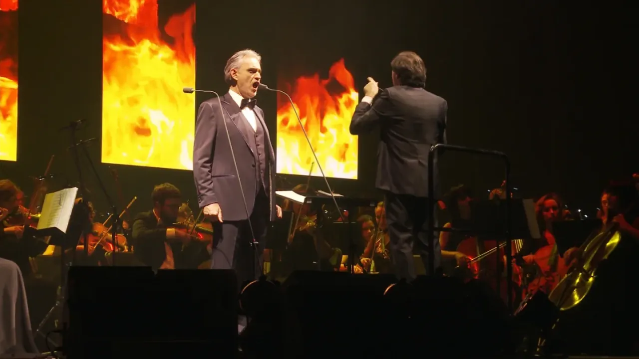 Andrea Bocelli śpiewa na koncercie, gdzie orkiestra gra na tle płomieni.