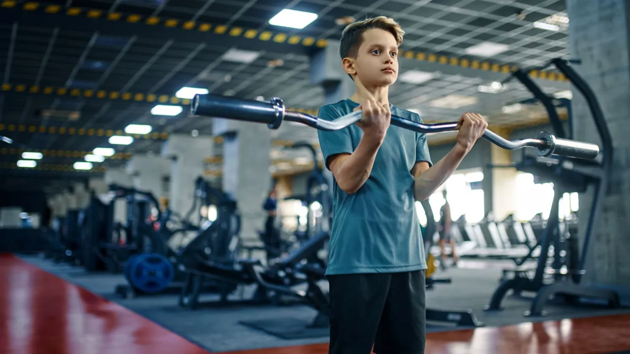 Chłopiec ćwiczy z gryfem. Od ilu lat można chodzić na siłownię? Odpowiedź zależy od wielu czynników, ale już od najmłodszych lat można zacząć przygodę ze sportem.