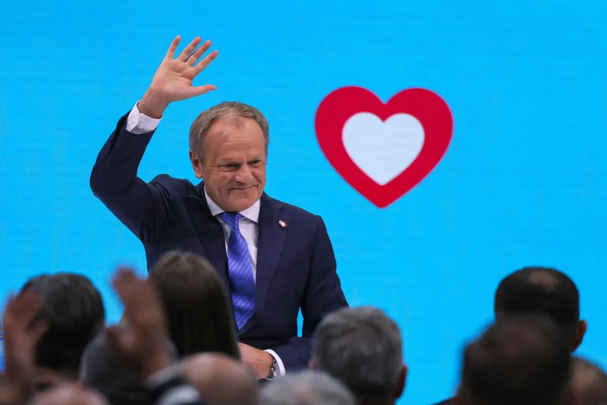 Donald Tusk macha do tłumu na tle niebieskiego ekranu z czerwonym sercem.