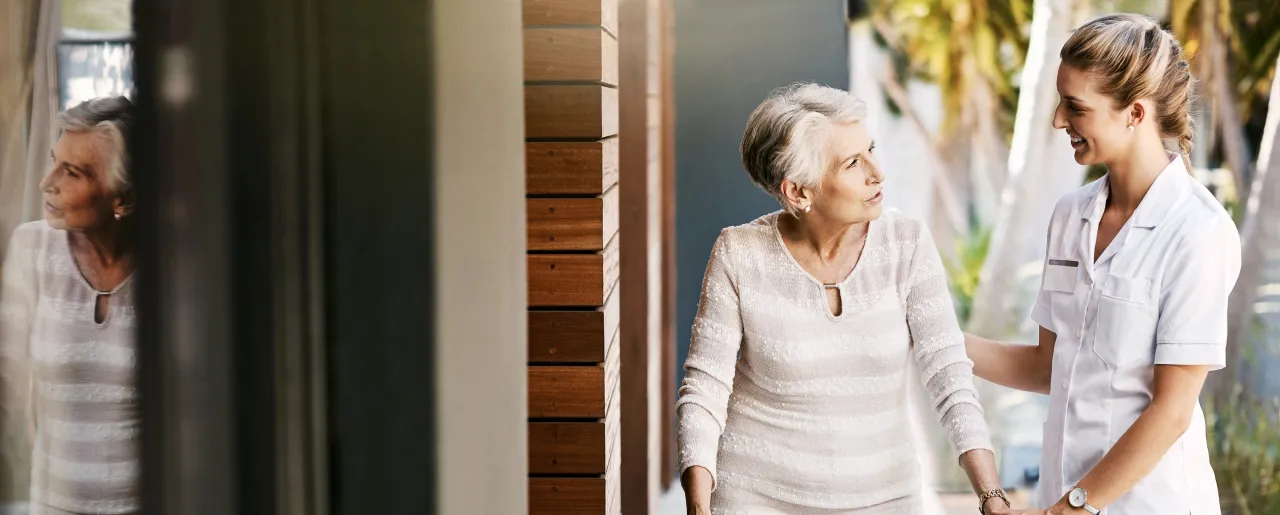 Uśmiechnięta opiekunka pomaga starszej kobiecie. Dowiedz się, ile kosztuje opieka nad starszą osobą za godzinę.