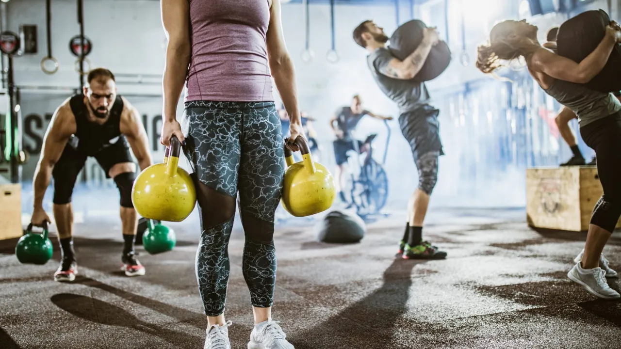 Intensywny trening obwodowy co to? Ludzie podnoszą ciężary, wykonują ćwiczenia z kettlebellami i workami treningowymi w siłowni.