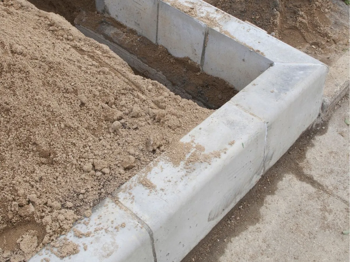 Piaskowy fundament pod szklarnię, wykonany z betonowych element&oacute;w i piasku.