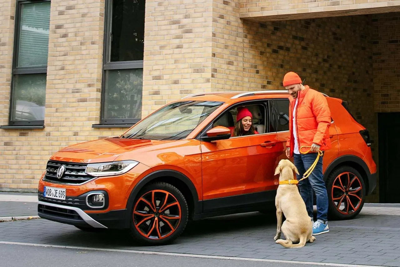 Pomarańczowy Volkswagen T-Cross, idealny samoch&oacute;d dla kobiety, z uśmiechniętą kierowczynią i mężczyzną wyprowadzającym psa.